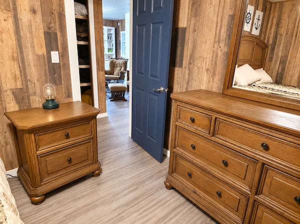A hallway with wooden flooring, a blue door, a wooden nightstand with a lamp, and a dresser with drawers.