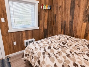 A bedroom with a bed, wooden walls, a window with blinds, a wall-mounted coat rack, and a heater.