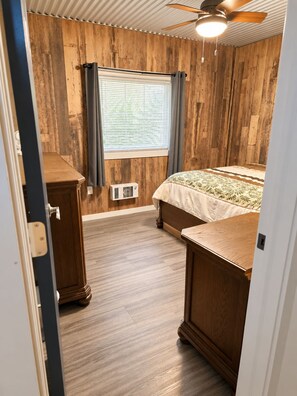A bedroom with a wooden floor, a bed with a patterned bedspread, a ceiling fan, and a window with curtains.