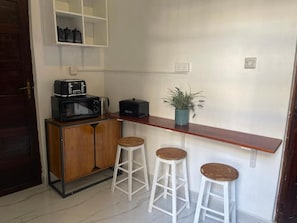A kitchenette with a microwave, toaster, and coffee maker on a wooden cabinet. There are three white stools and a potted plant on a shelf. - NASHU HOUSE STONE TOWN (Zanzibar)