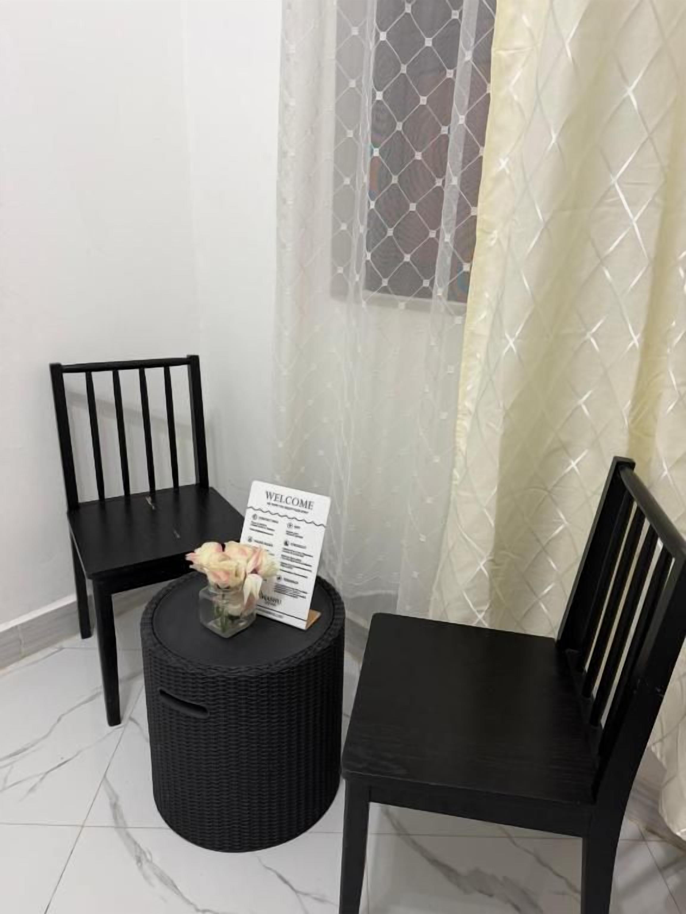 Una esquina de una habitación con dos sillas negras, una mesa redonda negra con un cartel de bienvenida y una cortina translúcida.