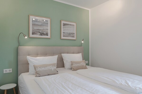 A bedroom with a bed, two pillows, a headboard, two wall-mounted lamps, and two framed pictures. - Nele - Wenningstedt-Braderup (Sylt) (Wenningstedt-Braderup (Sylt))
