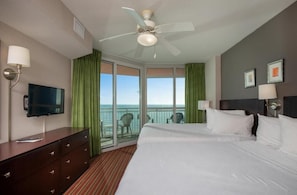 Ein Hotelzimmer mit Bett, Fernseher, Schreibtisch, Deckenventilator und einem Balkon mit Meerblick.