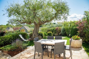 An outdoor patio with a table and chairs, surrounded by a garden with various plants and a large tree. - New Listing (Puglia)