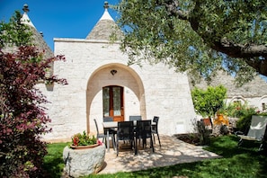 A stone building with a central archway, a dining table with chairs, and a garden with various plants. - New Listing (Puglia)