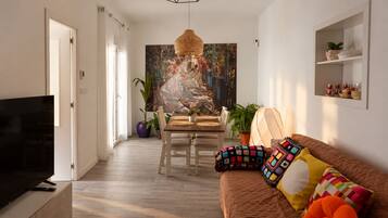 A modern living room with a television, a dining area with a table and chairs, and a sofa with colorful cushions.