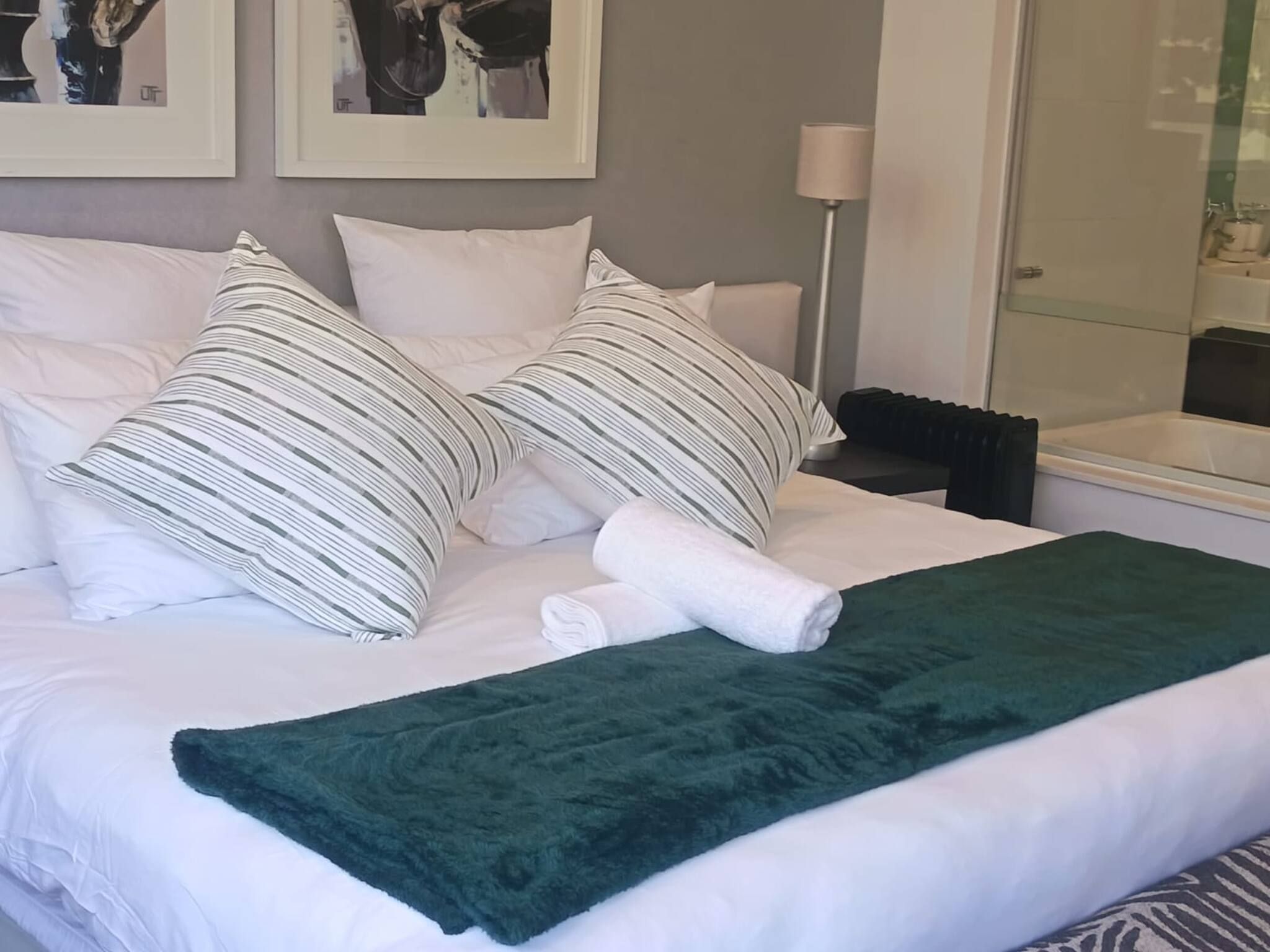 Una cama bien hecha con ropa blanca y almohadas a rayas, una manta verde y un cuadro colgado encima.