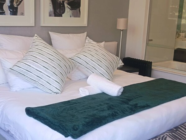 Cama bem arrumada com roupa de cama branca e almofadas listradas, um cobertor verde e um quadro em cima.