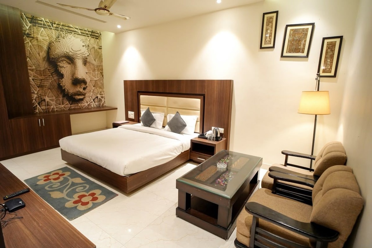 Una habitación de hotel con una cama, mesitas de noche, una mesa de centro, un sofá, una lámpara y obras de arte en las paredes.