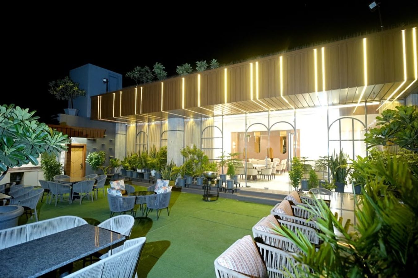 An outdoor seating area with tables and chairs, surrounded by potted plants and a building with illuminated vertical lines.