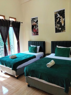 A hotel room with two beds, each with a green bedspread and white pillows, a window with curtains, and a wall with two framed artworks.