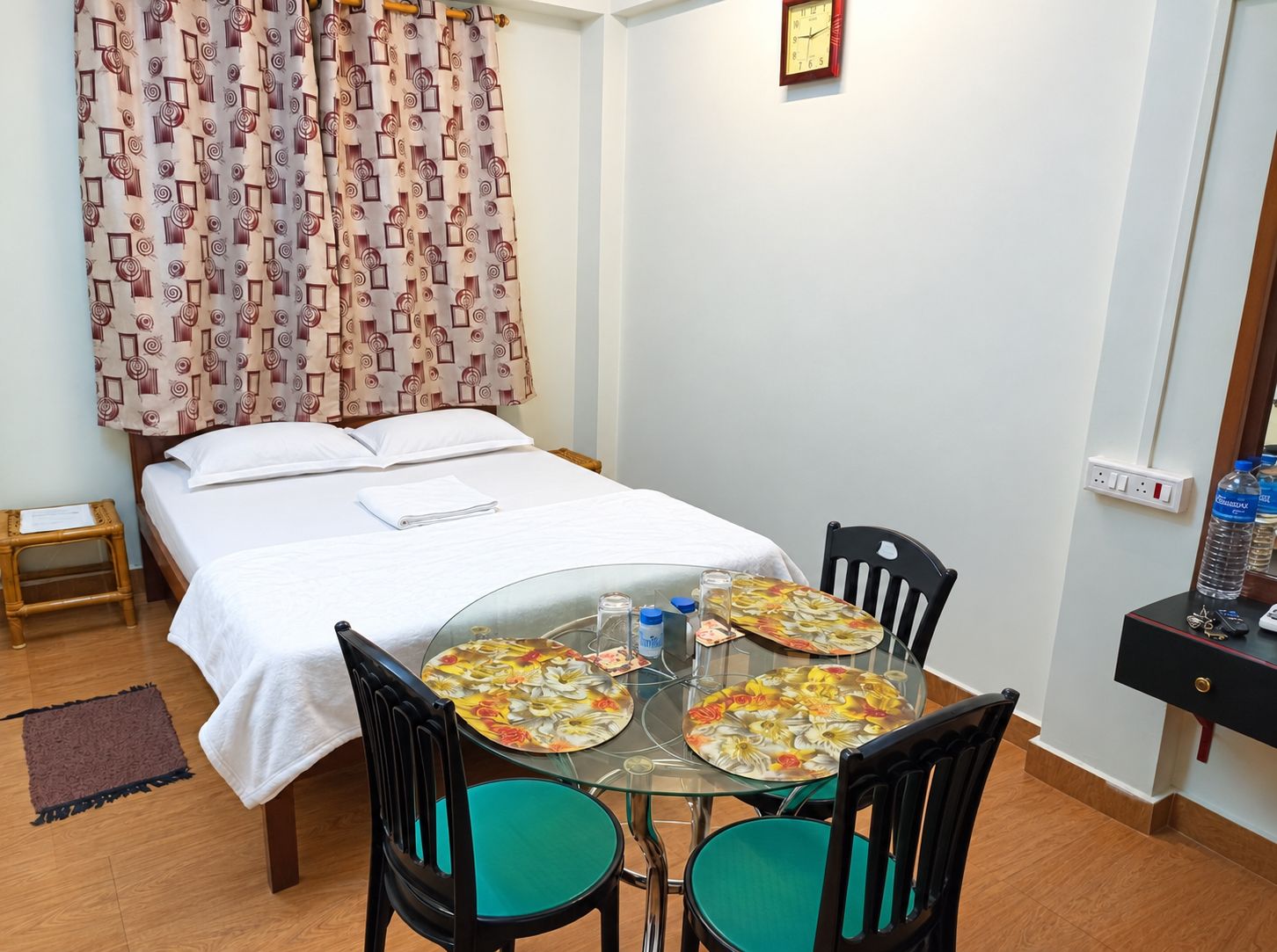A hotel room with a bed, a small table with chairs, a clock on the wall, and a power outlet.
