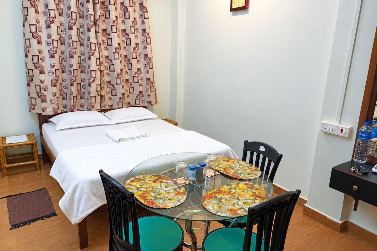 Una camera d'albergo con un letto, un tavolino con sedie, una sveglia al muro e una presa elettrica.