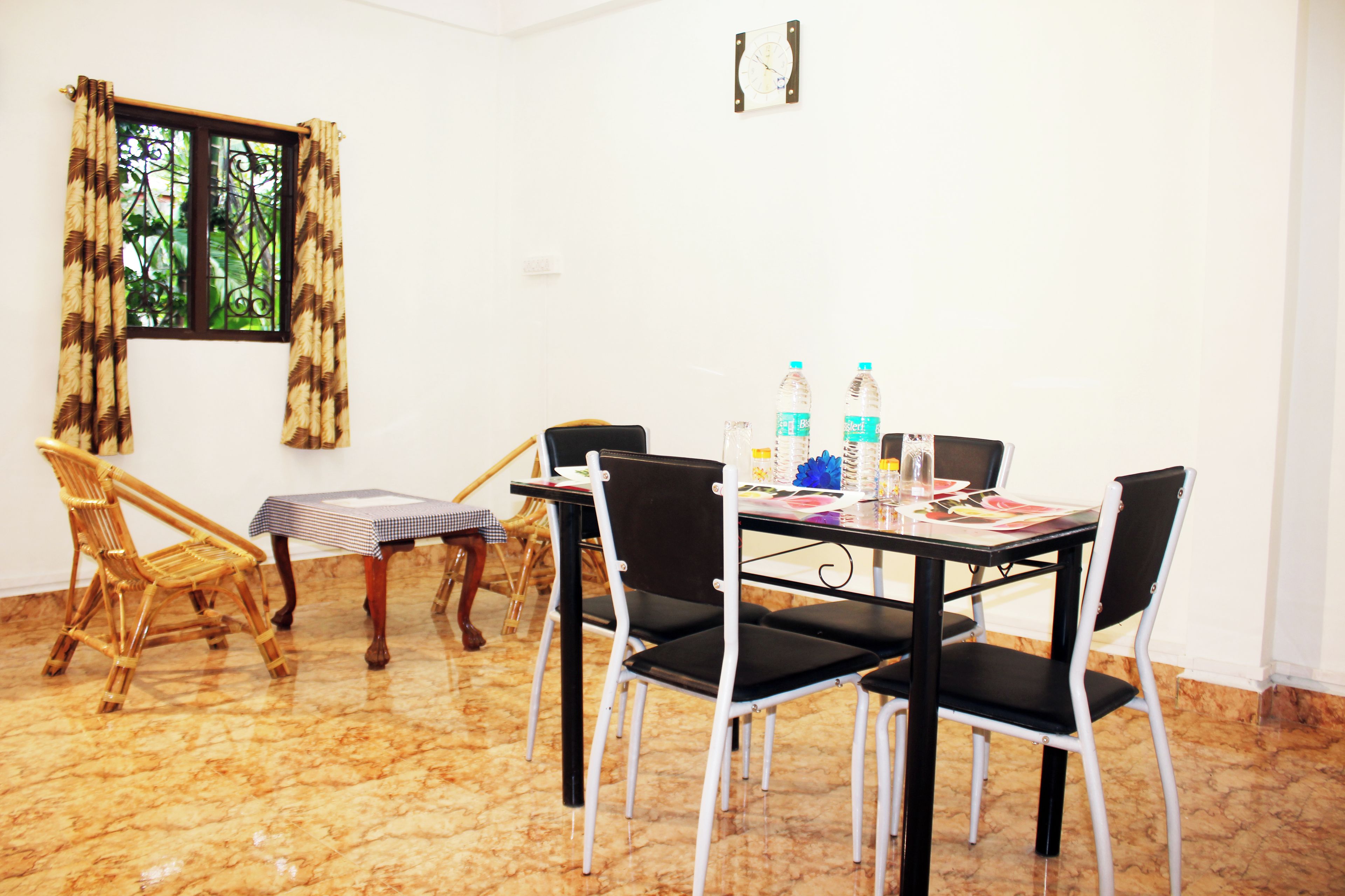 A dining area with a table set for four, surrounded by chairs, a window with curtains, and a clock on the wall.