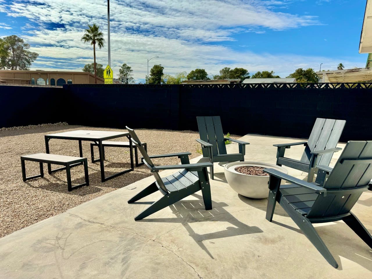 An outdoor seating area with Adirondack chairs and tables, a fire pit, and a view of the sky and trees.