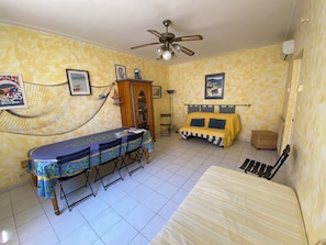 Quarto com cama, mesa e cadeiras, rede, ventilador de teto e estante de livros.