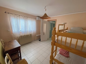 Quarto com cama de madeira, uma mesa pequena, uma cadeira, janela com cortinas leves e um ventilador de teto.