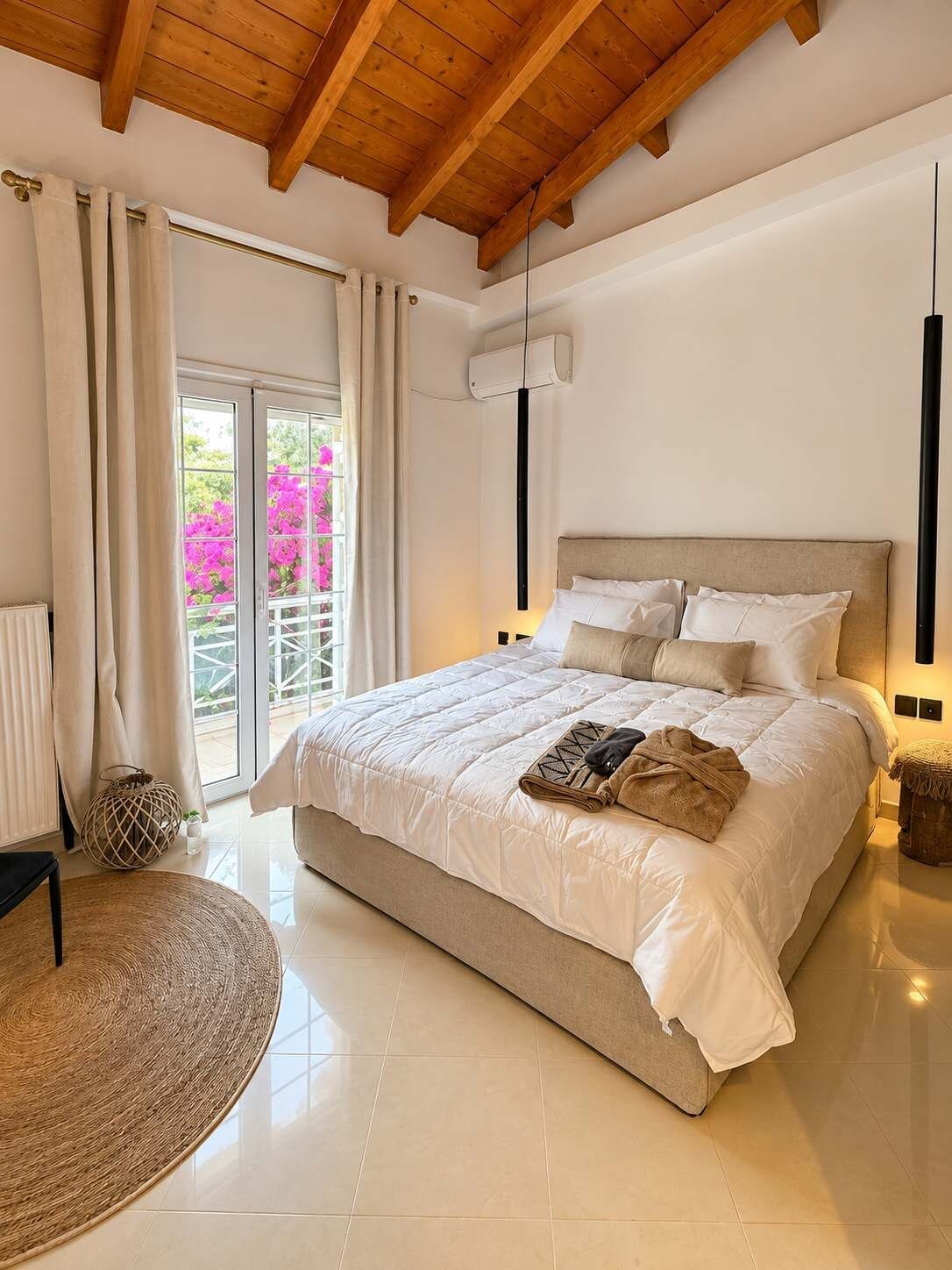 A bedroom with a large bed, a ceiling fan, a window with curtains, and a view of pink flowers outside.