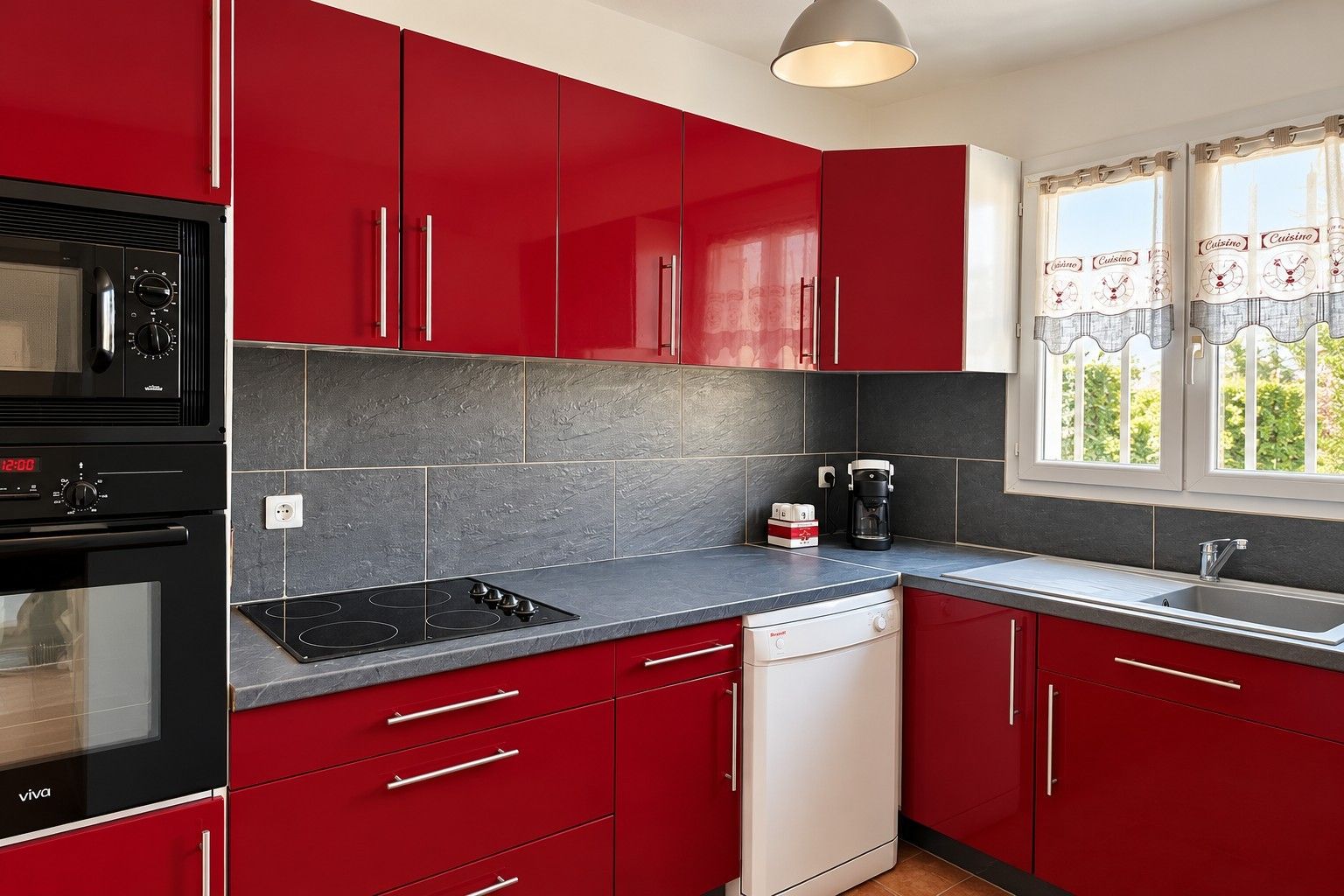 A kitchen with red cabinets, a black stove, a dishwasher, and a window with curtains.