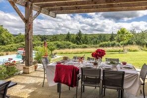 An outdoor dining area with a table set for a meal, surrounded by chairs, with a pool and greenery in the background. - New Listing (Milhac-de-Nontron)