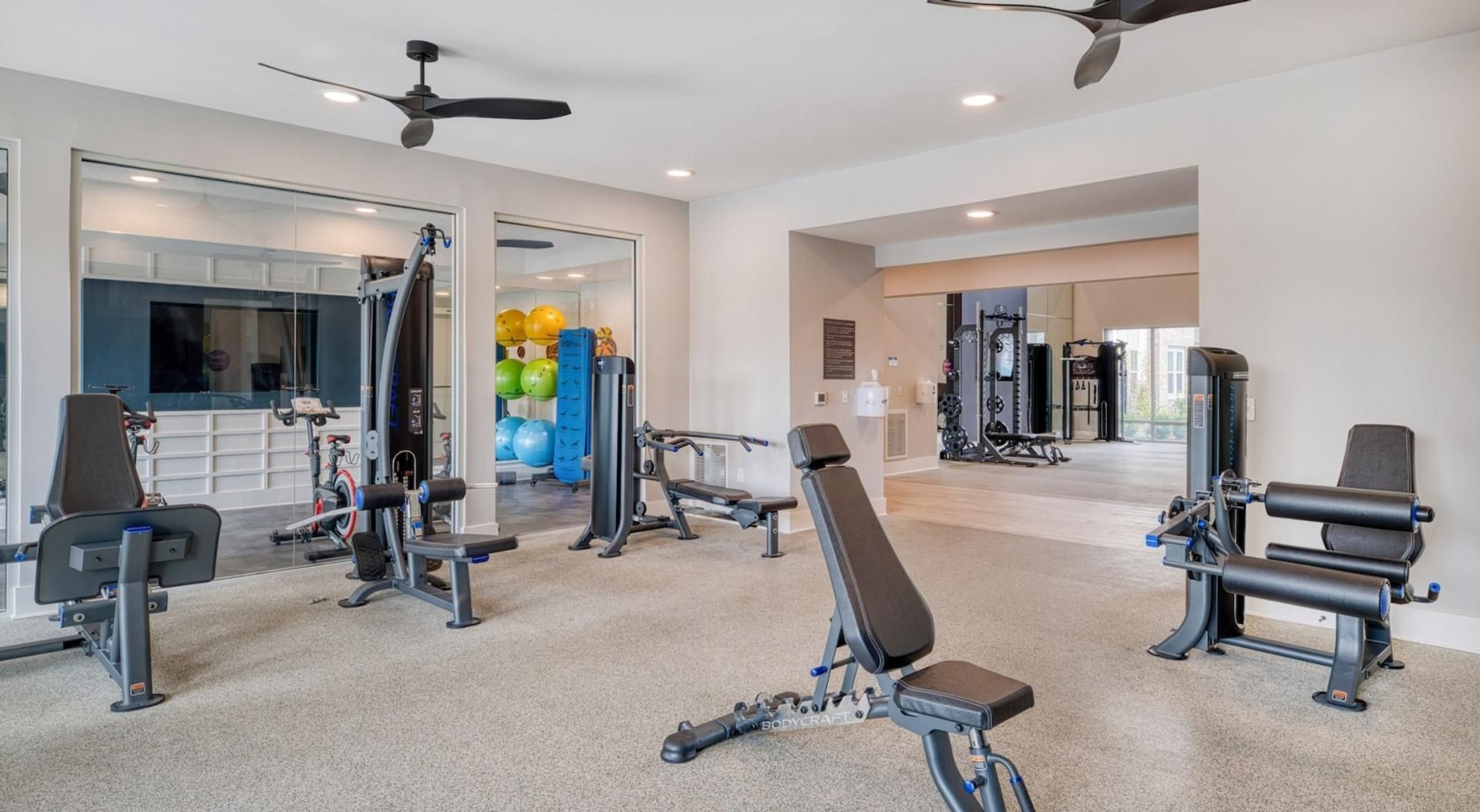 A gym with various exercise equipment, including weight machines and free weights, and a ceiling fan.