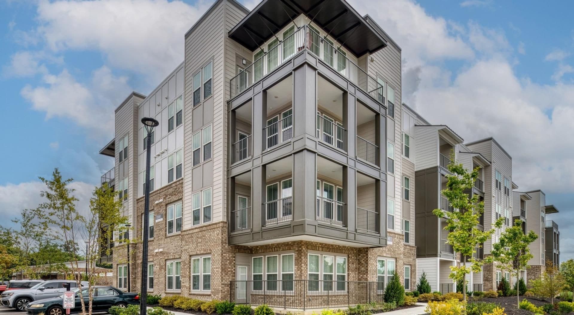 A multi-story residential building with a corner entrance, balconies, and a parking area with cars.
