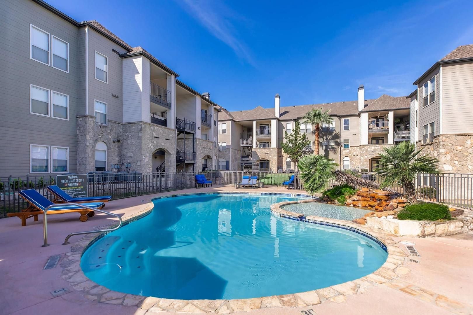 A swimming pool surrounded by apartment buildings with balconies, a stone wall, and lounge chairs.