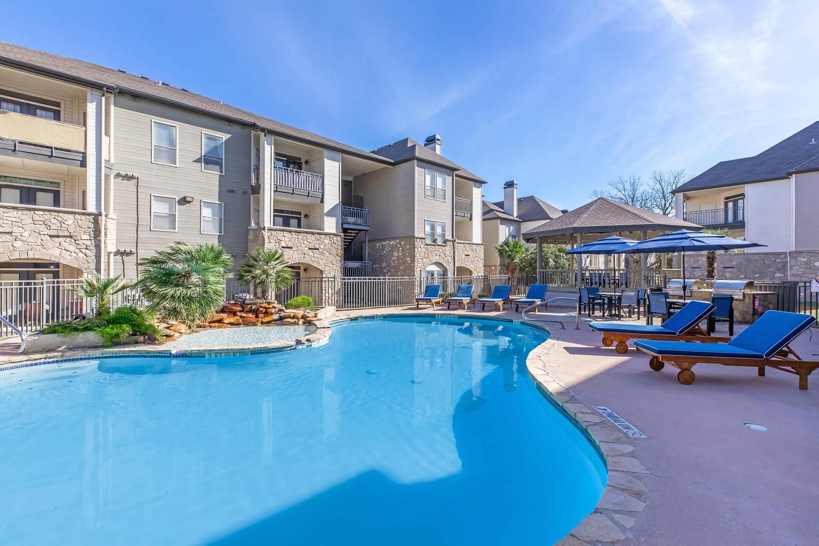 A swimming pool with lounge chairs and umbrellas, surrounded by multi-story buildings with balconies.