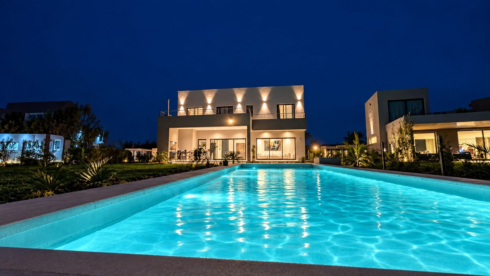 A modern building with a swimming pool in front, surrounded by well-lit landscaping.