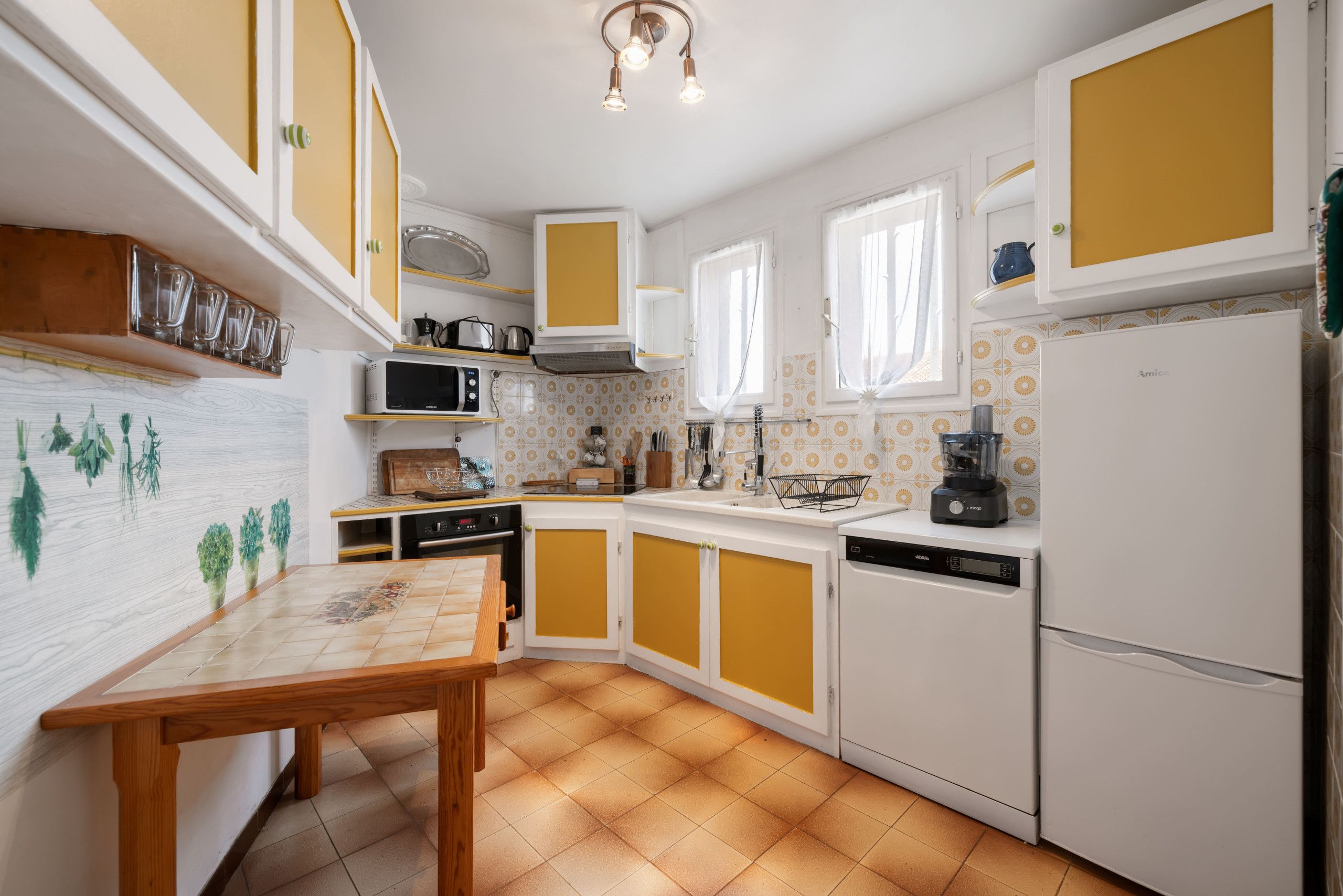 Une cuisine avec des meubles blancs et jaunes, une table en bois, un four à micro-ondes, une cuisinière et un réfrigérateur.