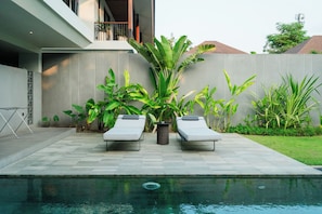 Un área de piscina exterior moderna con dos sillas de descanso y una variedad de plantas tropicales.