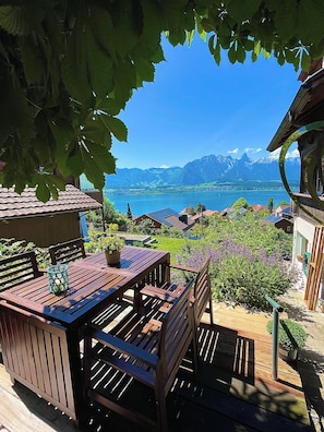 Terraza de madera con una mesa y sillas, con vista a un lago y montañas al fondo.