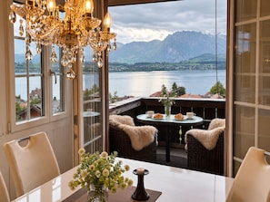 A dining area with a view of a lake and mountains, featuring a chandelier, wicker furniture, and a vase with flowers. - Mon Bijou Luxury Chalet au Lac – Wake Up to Lake Thun & Alps Views – 95 m² (Hilterfingen)