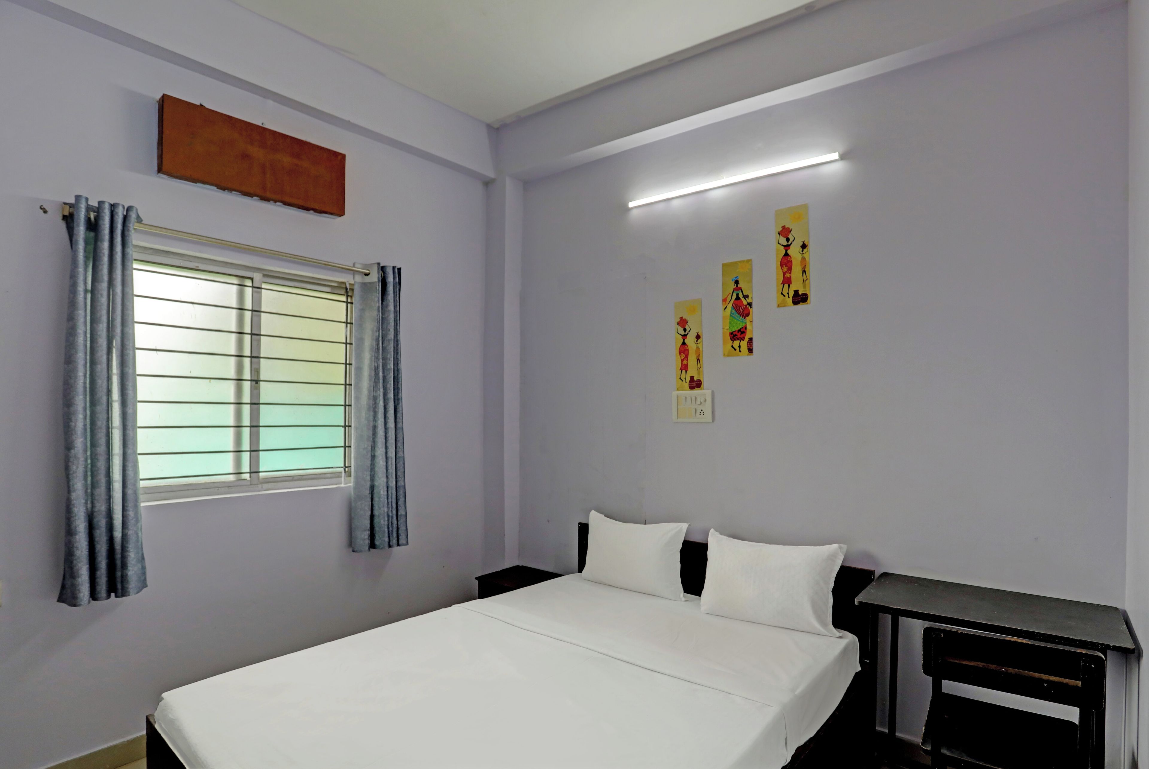 Ein Schlafzimmer mit einem Bett, einem Fenster mit Vorhängen, einem Nachttisch und Wandbildern.