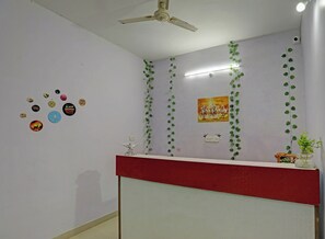 A reception area with a red counter, a ceiling fan, wall-mounted light, and decorative plants. - Hotel O 815596 Hotel Sambodhi Palace (Bhopal)