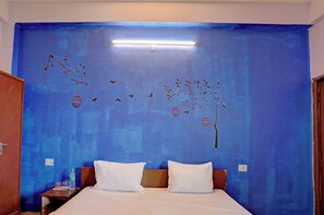 A room with a bed, two pillows, a nightstand with a bottle, and a wall with a blue mural. - Hotel O 815596 Hotel Sambodhi Palace (Bhopal)