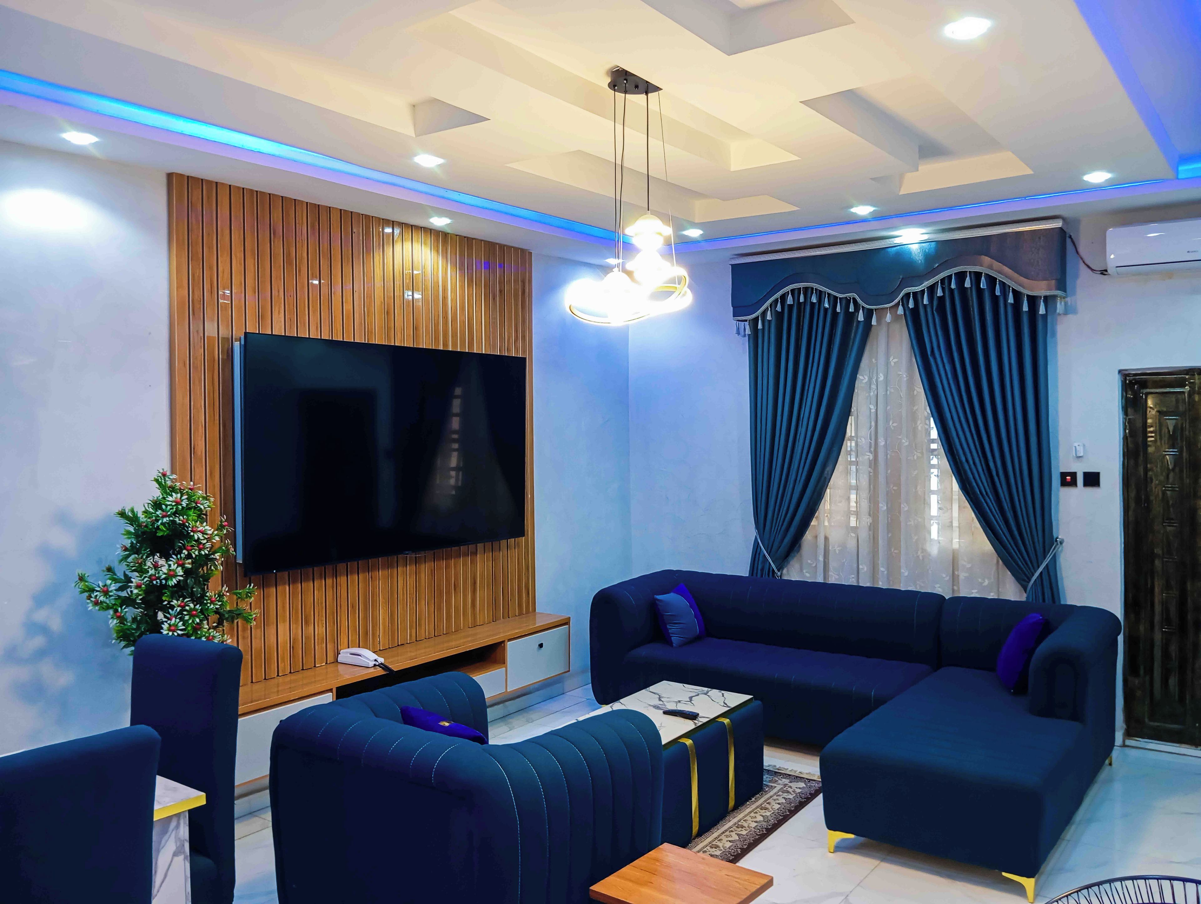 A modern living room with a flat-screen TV, dark blue sofas, a wooden panel wall, and a ceiling with recessed lighting.
