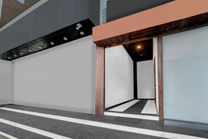 Entrada de un edificio moderno con una rampa de hormigón que lleva a una puerta, un alero de madera sobre la puerta y una rejilla de ventilación en la pared.
