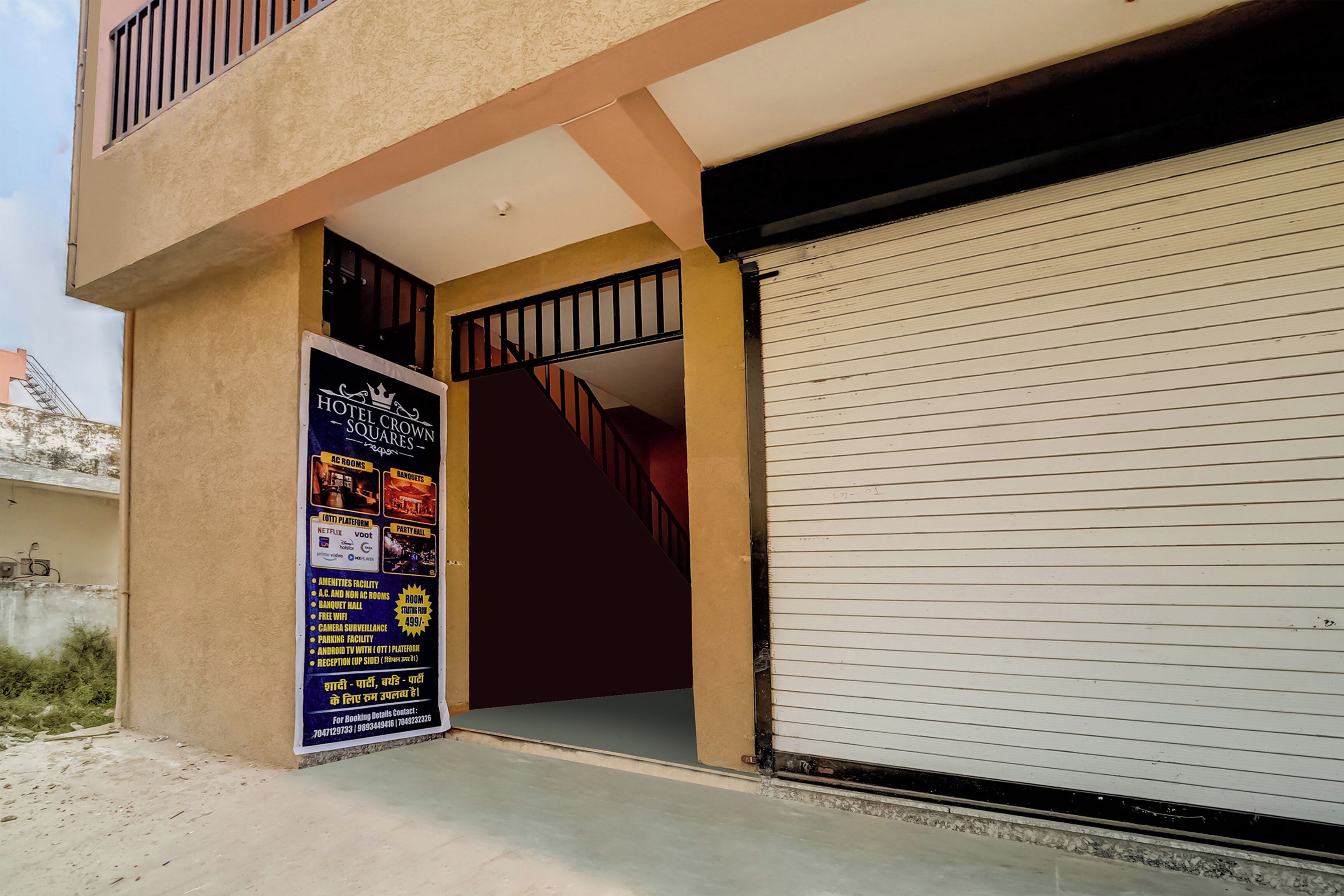 A hotel entrance with a closed shutter, a staircase leading to an upper floor, and a signboard advertising amenities like AC rooms, banquet hall, and free WiFi.