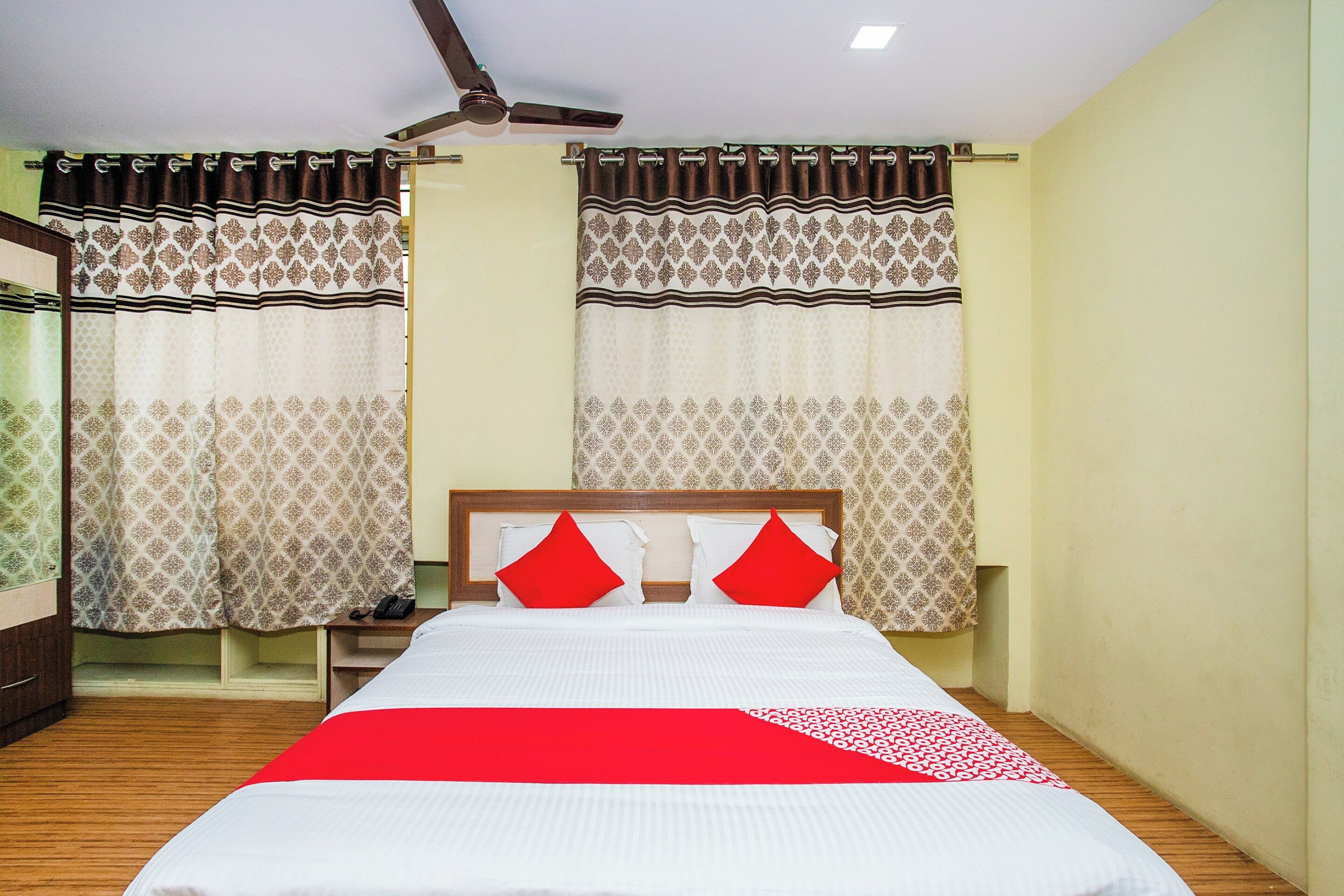 A hotel room with a bed, curtains, a ceiling fan, and a bedside telephone.