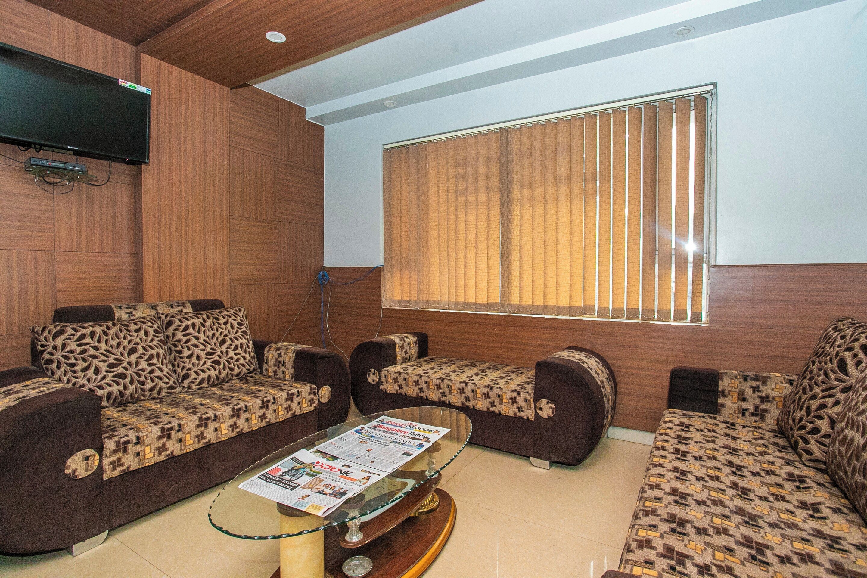 A living room with patterned sofas, a coffee table with newspapers, and a television mounted on the wall.