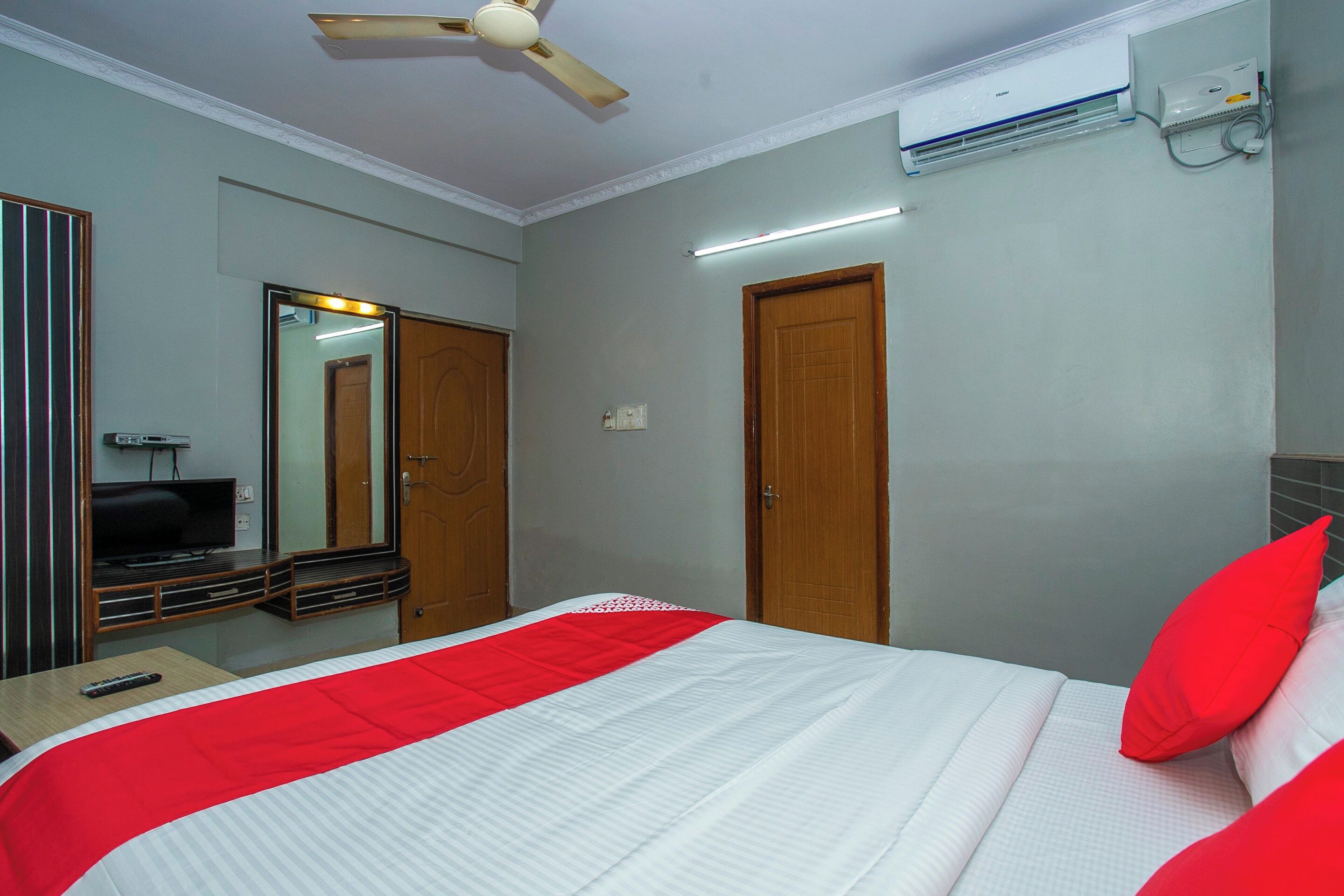 A hotel room with a bed, a ceiling fan, an air conditioning unit, a mirror, and a television.