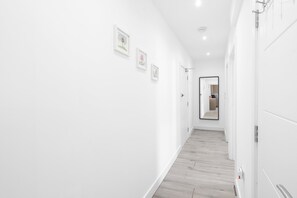 Un couloir lumineux, aux murs blancs et au plancher en bois, qui mène à une pièce dotée d’un miroir et d’une porte.