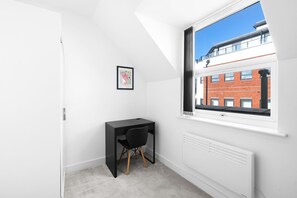 Une petite pièce blanche, avec un bureau et une chaise noirs, un tableau encadré au mur et une fenêtre donnant sur un bâtiment en briques rouges.