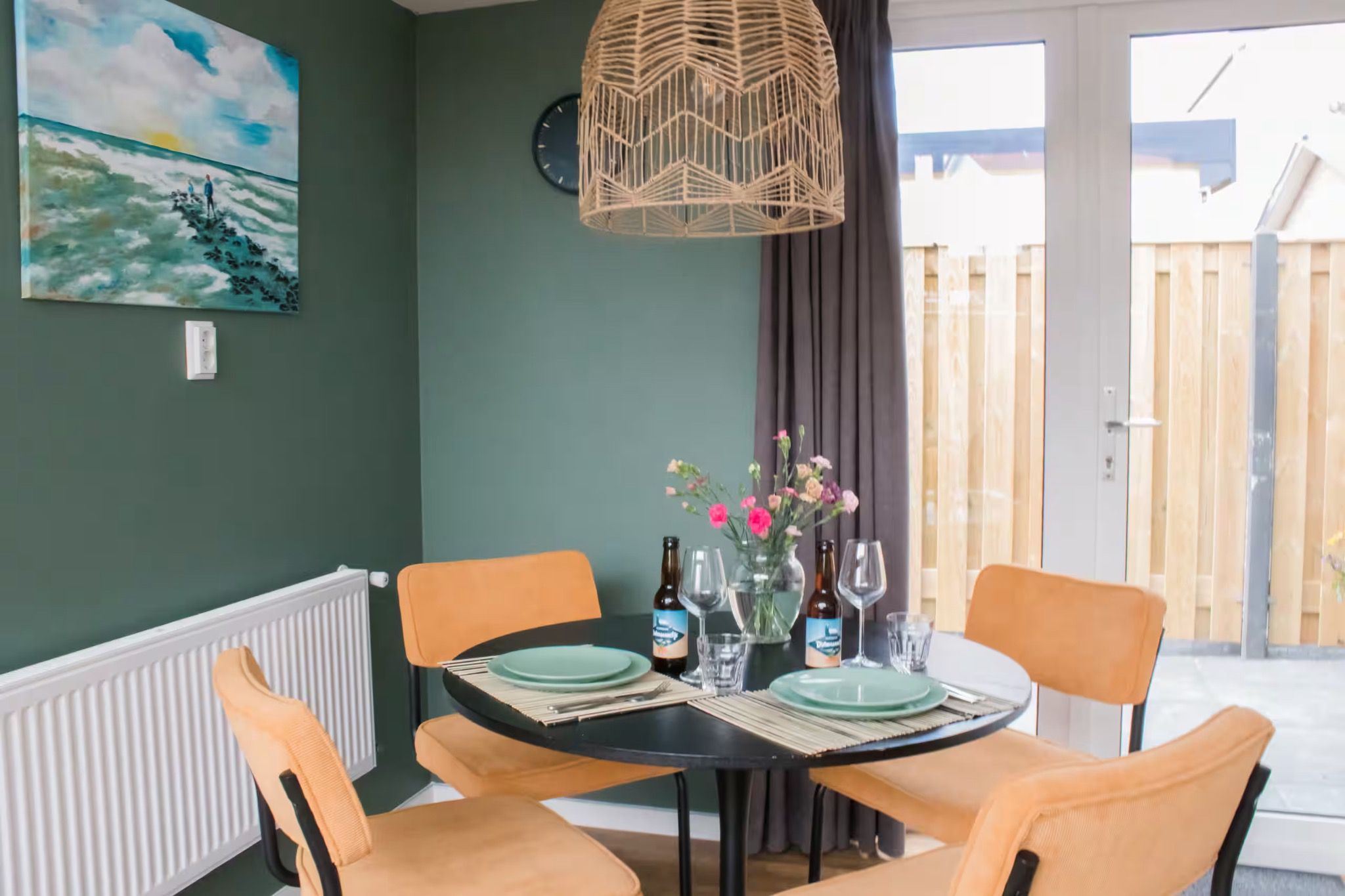 A dining area with a round table set for two, featuring a woven light fixture above, a clock on the wall, and a view of a wooden fence outside.