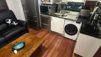 A compact kitchen with a refrigerator, microwave, stove, and sink. There is a wooden table with a bowl and a black sofa in the living area.