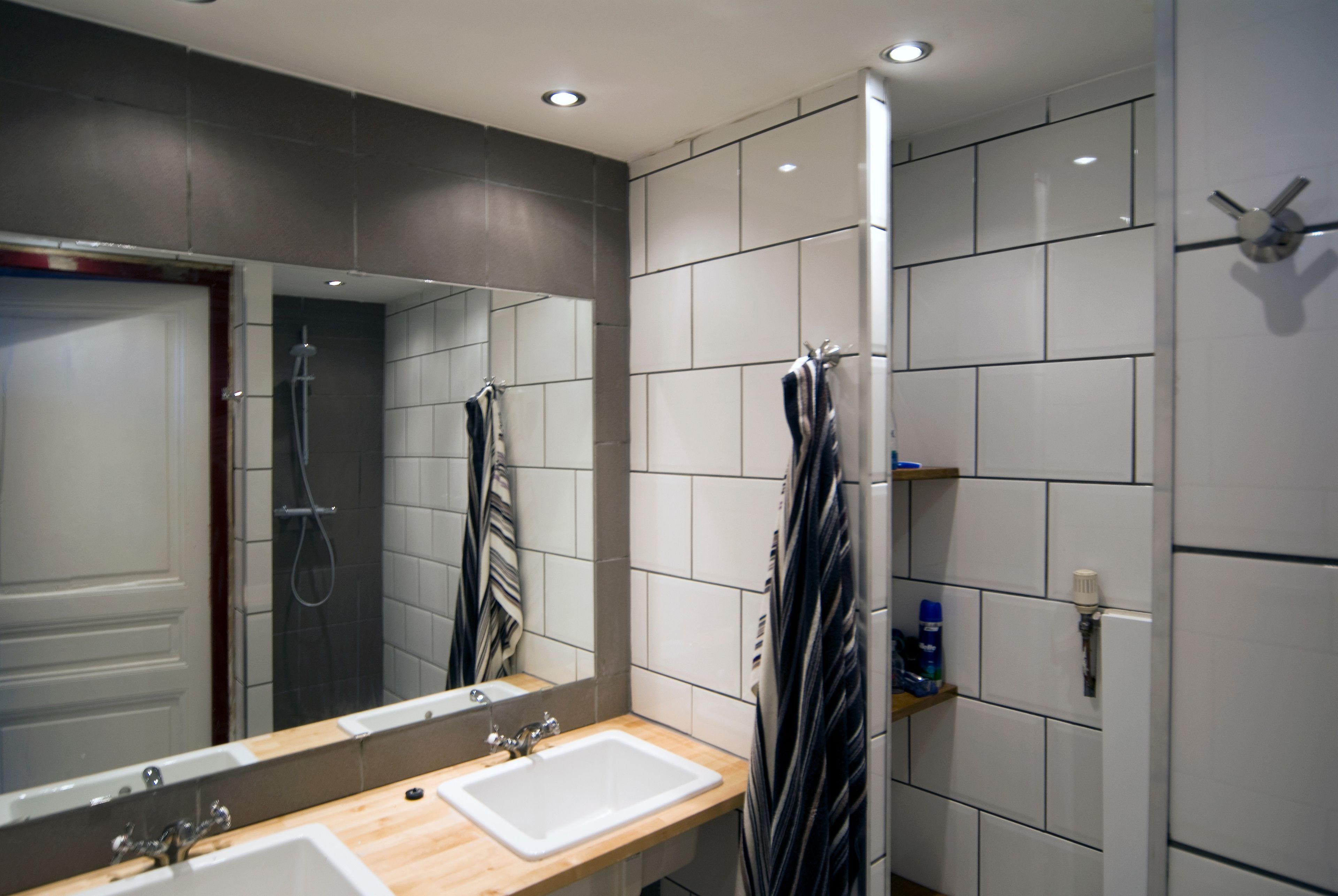 A modern bathroom with a large mirror, two sinks, a shower, and tiled walls.