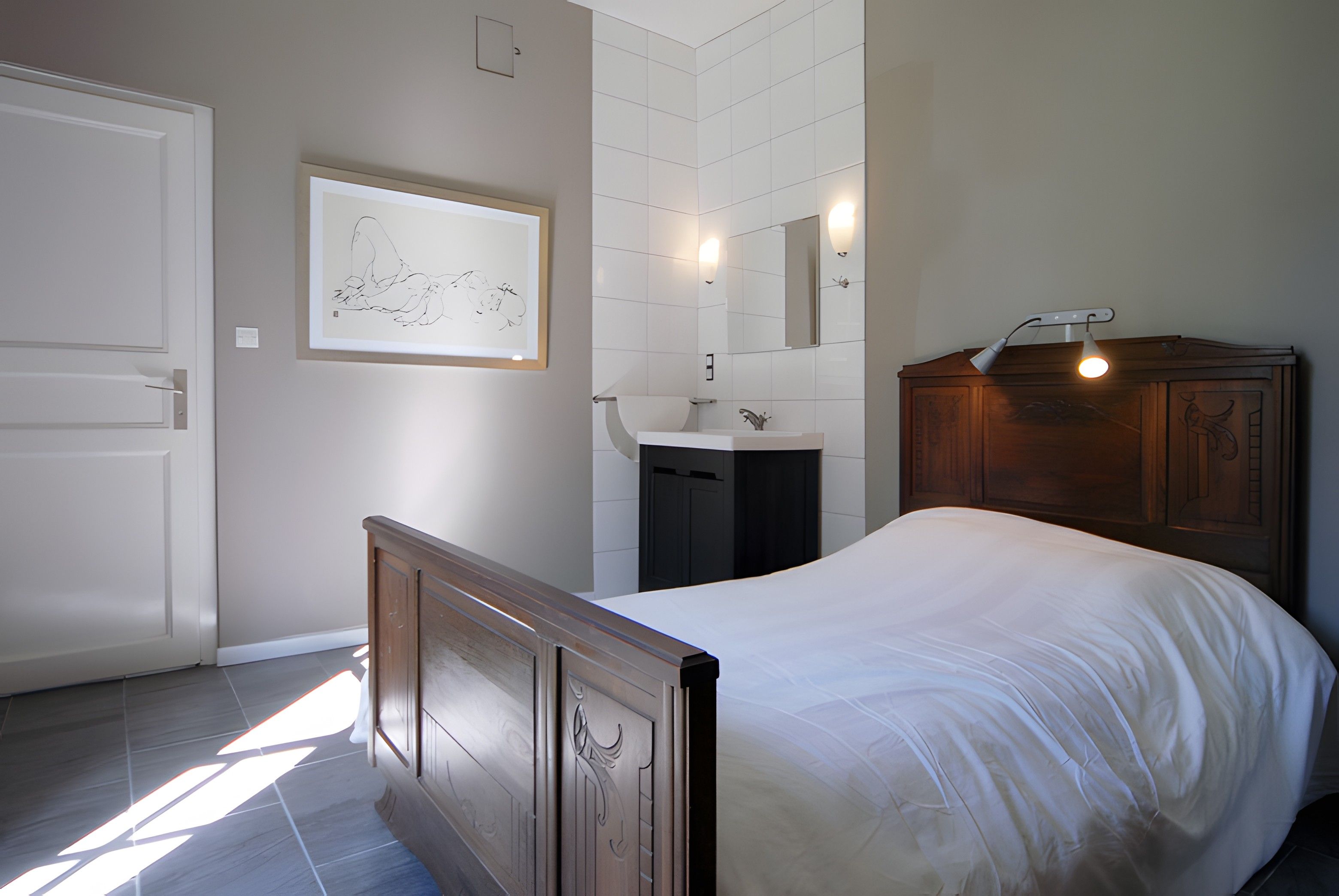 A bedroom with a bed, a wooden wardrobe, a bathroom with a sink and mirror, and a framed artwork on the wall.