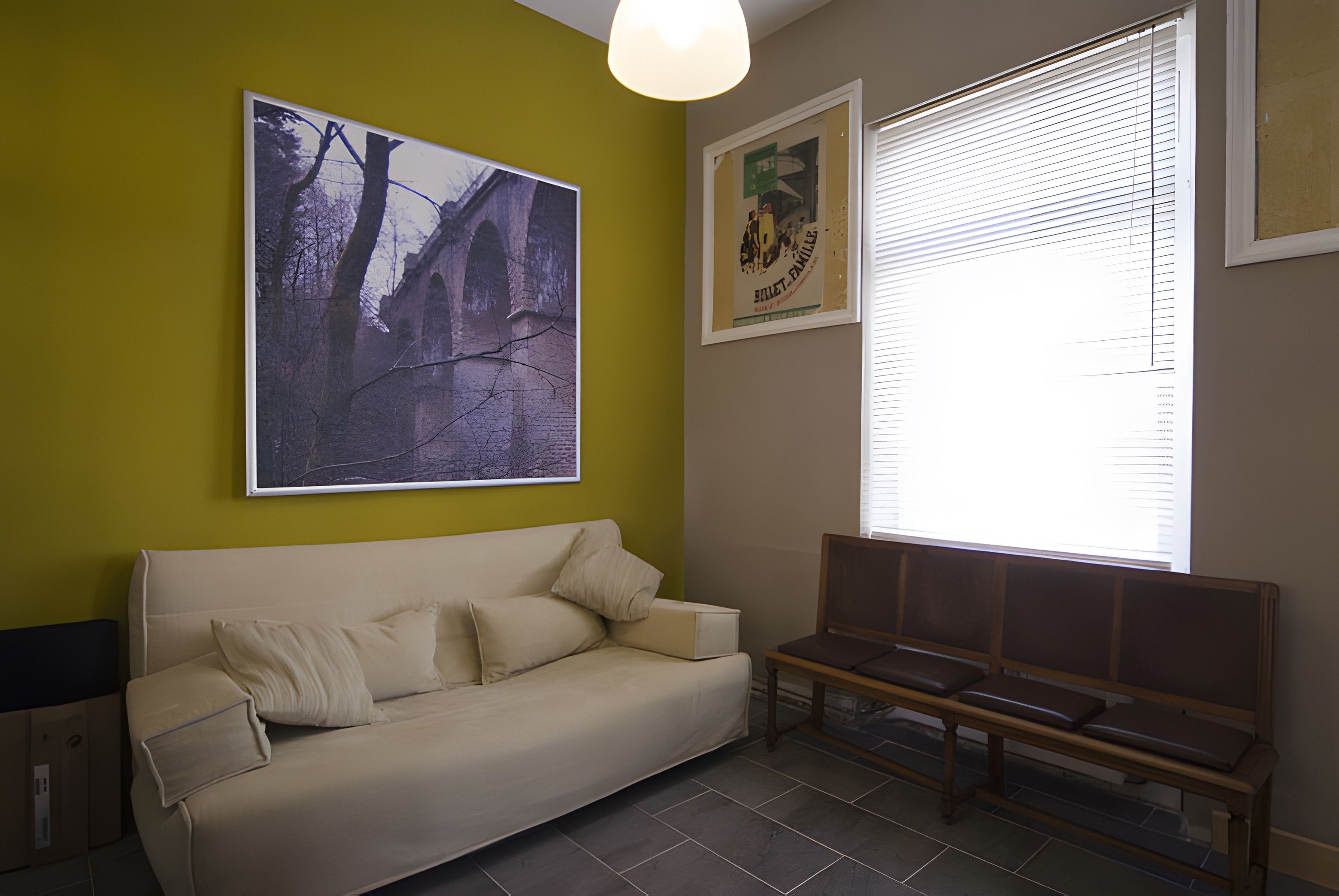 Ein Zimmer mit einer Couch, einer Bank, einem Fenster mit Jalousien und zwei gerahmten Bildern an der Wand.
