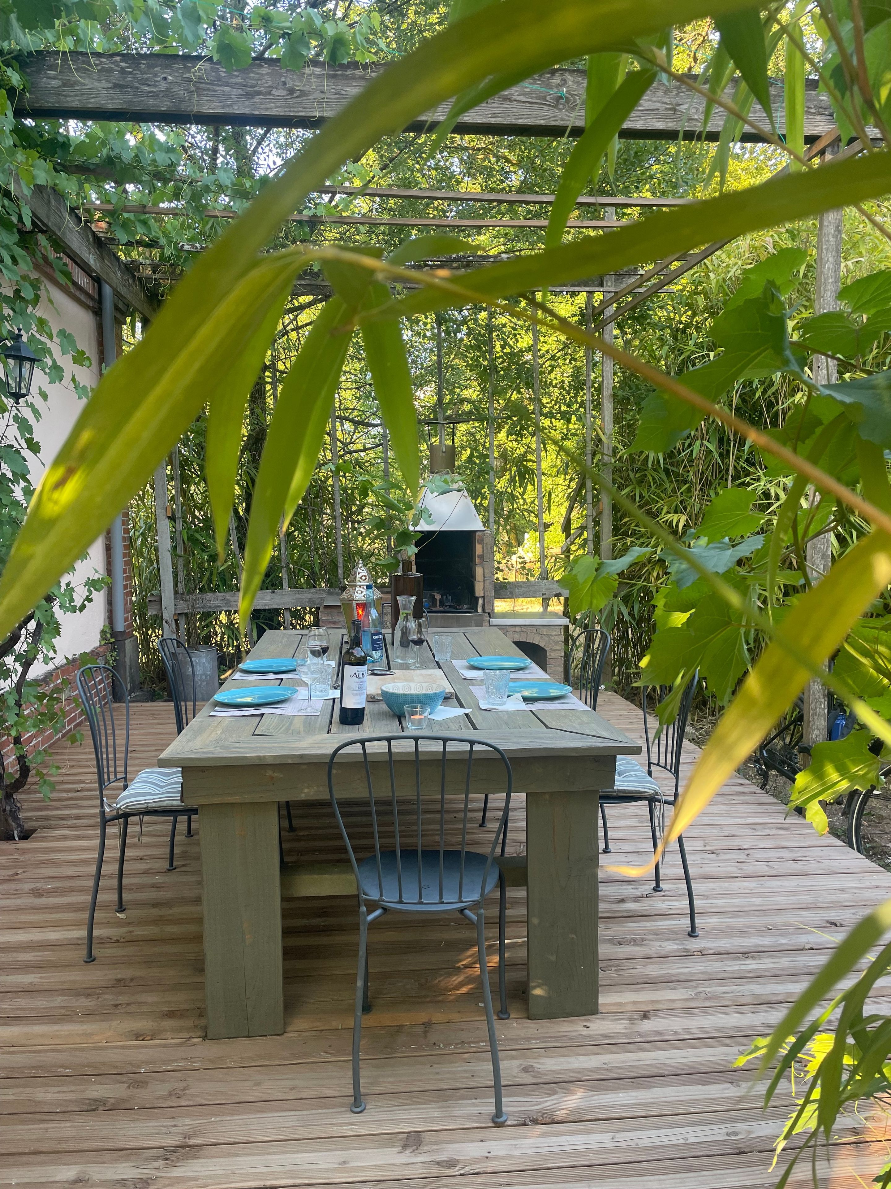 Una terraza de madera al aire libre con una mesa lista para comer, rodeada de follaje y una estructura de pérgola.